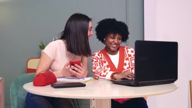Birlikte çalışırken bilgisayar ve akıllı telefon kullanarak masada oturan gülümseyen öğrenciler. Samimi ve dost canlısı bir öğrenme alanında bir video çağrısına ya da çevrimiçi aktiviteye katılıyorlar.. 