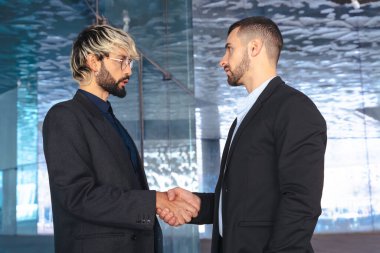 Modern cam bir ofiste takım elbiseli iki profesyonel işadamı el sıkışıyor. İş anlaşması, anlaşma ya da çağdaş tasarım ile kurumsal bir iç mekanda ortaklık. 