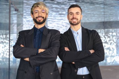 Takım elbiseli iki kendine güvenen profesyonel erkek kollarını kavuşturup modern bir ofisin içinde kameraya gülümsüyor. Şirket başarısı, takım çalışması ve iş ortamında liderlik.. 