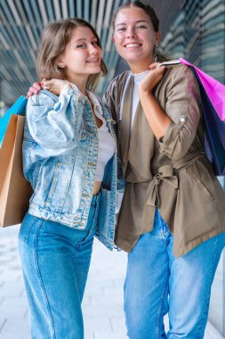 Renkli alışveriş torbaları taşıyan ve alışveriş merkezinde başarılı bir günün ardından gülümseyen iki beyaz kadın. Modern bir binanın dışında duruyorlar, birlikte eğleniyorlar stil ve neşe içinde. Dikey
