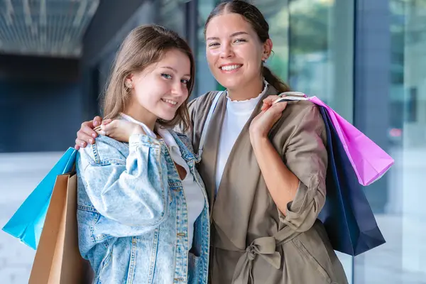 Renkli alışveriş torbaları taşıyan ve alışveriş merkezinde başarılı bir günün ardından gülümseyen iki beyaz kadın. Modern bir binanın önünde duruyorlar, birlikte eğleniyorlar stil ve neşeyle. 