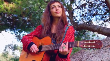 Genç bayan müzisyen, sakin bir açık hava parkında akustik gitarla şarkı söylüyor. Doğa tarafından çevrelenmiş, tutku, yaratıcılık ve duyguyu gündelik bir ortamda müzik yoluyla ifade eder.. 
