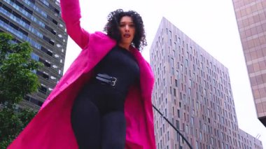 Pembe palto giyen kıvırcık saçlı zarif dansçı şehir ortamında hareketini ve enerjisini ifade ediyor. Modern moda, çağdaş dans ve sanatsal performans şehrin arka planına karşı..