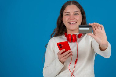 Beyaz kazaklı ve kırmızı kulaklıklı mutlu genç bir kadın kameraya bir kredi kartı gösterirken gülümsüyor ve elinde kırmızı bir akıllı telefon tutuyor, mavi arka planda, online alışverişi sembolize ediyor. Boşluğu kopyala