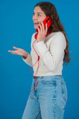 Kırmızı bir akıllı telefondan konuşan neşeli genç bir kadının dikey görüntüsü. Elini göğsüne koyup gülüyor. Parlak mavi bir arka plana yaslanıyor. Sıradan bir anda.. 