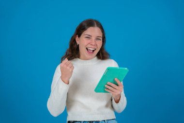 Beyaz kazaklı mutlu genç bir kadın yeşil bir tablet tutarken yumruğunu kaldırıyor ve kameraya gülümsüyor. Mavi arka plan, başarıyı, neşeyi ve dijital hayatı simgeliyor.. 