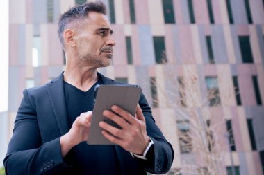 İş adamı, modern bir şehirde gözünü kaçırırken açık havada bir tablet kullanıyor. Siyah bir takım elbise ve şık bir bant giyiyor. Teknoloji, odaklanma, hareketlilik ve dijital mesleki yaşam tarzı kavramı. 