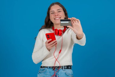 Beyaz kazaklı ve kırmızı kulaklıklı mutlu genç bir kadın kameraya bir kredi kartı gösterirken gülümsüyor ve elinde kırmızı bir akıllı telefon tutuyor, mavi arka planda online alışverişi sembolize ediyor.. 
