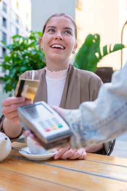 Açık bir kafede temassız teknoloji kullanan beyaz bir kadın kredi kartıyla ödeme yaparken gülümsüyor. Rahat, yaşam tarzı ve modern finansal alışkanlıkları yansıtan sıradan bir kentsel an. Dikey