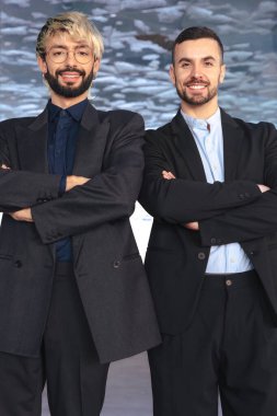 Takım elbiseli iki kendine güvenen profesyonel erkek kollarını kavuşturup modern bir ofisin içinde kameraya gülümsüyor. Şirket başarısı, takım çalışması ve iş ortamında liderlik. Dikey