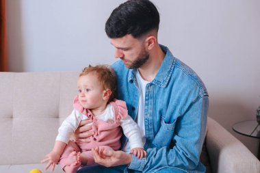 Kot gömlekli genç baba kanepeye otururken bebeğini nazikçe tutuyor. Bebek, evde sakin ve samimi bir ebeveynlik anını yakalamak için tuhaf bir şekilde uzağa bakıyor.. 