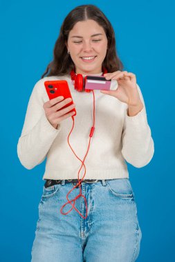 Beyaz kazaklı ve kırmızı kulaklıklı mutlu genç bir kadın kameraya bir kredi kartı gösterirken gülümsüyor ve elinde kırmızı bir akıllı telefon tutuyor, mavi arka planda, online alışverişi sembolize ediyor. Dikey