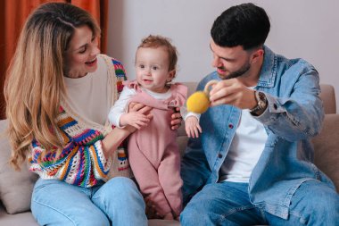 Küçük kızlarıyla oyuncak bir topla oynarken koltukta oturan gülümseyen ebeveynler. Aşk, aile bağları, erken gelişim ve ebeveynlik gibi konular için ideal ev ortamları. 