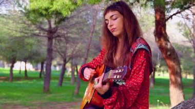 Genç bayan müzisyen, sakin bir açık hava parkında akustik gitarla şarkı söylüyor. Doğa tarafından çevrelenmiş, tutku, yaratıcılık ve duyguyu gündelik bir ortamda müzik yoluyla ifade eder.. 