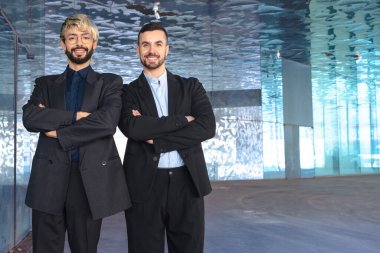Takım elbiseli iki kendine güvenen profesyonel erkek kollarını kavuşturup modern bir ofisin içinde kameraya gülümsüyor. Şirket başarısı, takım çalışması ve iş ortamında liderlik. Boşluğu kopyala