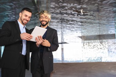 Resmi takım elbiseli iki genç işadamı, bir tablete bakıp gülümserken aynı zamanda yansıtıcı yüzeyleri ve profesyonel atmosferi olan şık, modern bir ofiste birlikte çalışıyorlar. Boşluğu kopyala