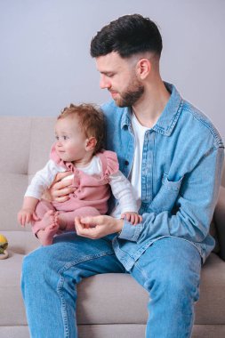 Kot gömlekli genç baba koltuğunda otururken bebeğini nazikçe tutuyor. Bebek, evde sakin ve samimi bir ebeveynlik anını yakalamak için tuhaf bir şekilde uzağa bakıyor.. 