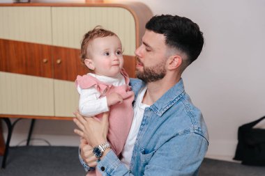 Kot gömlekli genç baba kanepeye otururken bebeğini nazikçe tutuyor. Bebek, evde sakin ve samimi bir ebeveynlik anını yakalamak için tuhaf bir şekilde uzağa bakıyor.. 