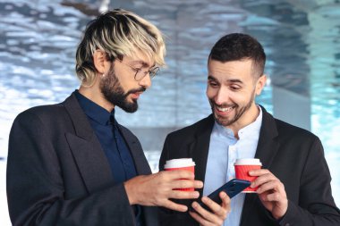 Takım elbiseli iş adamları modern bir ofiste birlikte akıllı telefona bakarken kahveden hoşlanırlar. Günlük ve dinamik ortam, takım çalışması, iletişim ve teknoloji gibi konular için ideal. 