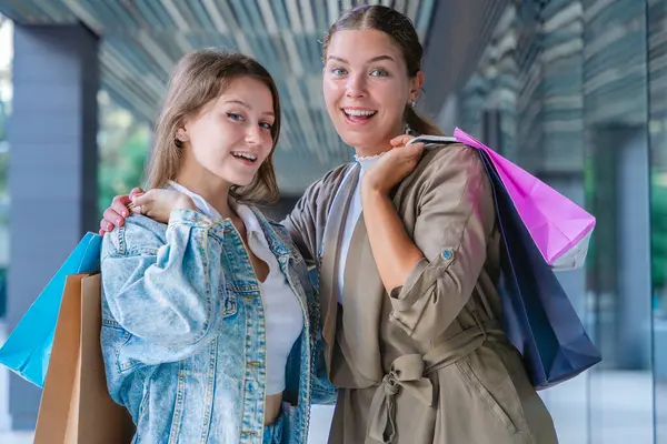Renkli alışveriş torbaları taşıyan ve alışveriş merkezinde başarılı bir günün ardından gülümseyen iki beyaz kadın. Modern bir binanın önünde duruyorlar, birlikte eğleniyorlar stil ve neşeyle. 
