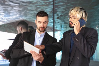 Modern bir ofiste iş birliği yapan takım elbiseli iki genç iş adamı. Biri telefonda konuşurken diğeri tablet kullanıyor, yansıtıcı camlar ve şık profesyonel bir atmosfer ile çevrili.. 