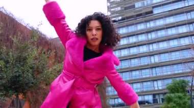 Pembe palto giyen kıvırcık saçlı zarif dansçı şehir ortamında hareketini ve enerjisini ifade ediyor. Modern moda, çağdaş dans ve sanatsal performans şehrin arka planına karşı..