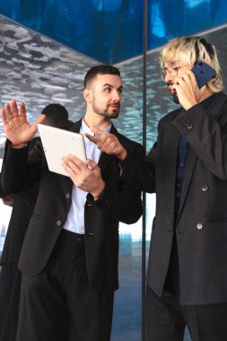 Modern bir ofiste iş birliği yapan takım elbiseli iki genç iş adamı. Biri telefonda konuşurken diğeri tablet kullanıyor, yansıtıcı camlar ve şık profesyonel bir atmosfer ile çevrili. Dikey