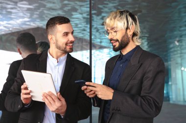 Resmi takım elbiseli iki genç işadamı, bir tablete bakıp gülümserken aynı zamanda şık, modern bir ofiste yansıtıcı yüzeylere ve profesyonel bir atmosfere sahipler.. 
