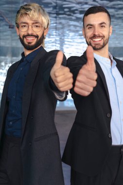 Resmi takım elbiseli iki genç iş adamı kameraya güvenle gülümsüyor, yansımaları ve şık iç tasarım unsurları olan modern cam bir ofiste başparmak hareketi yapıyorlar.. 