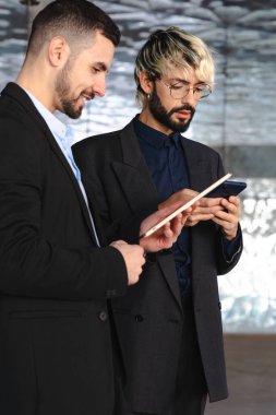 Takım elbiseli iki genç işadamı, modern, şık bir ofis alanında yansıtıcı cam duvarlar ve profesyonel, teknoloji odaklı bir atmosferle işbirliği yaparken dijital tabletlere odaklandılar. dikey