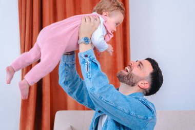 Kot ceketli genç baba, pembe tulumlu, gülümseyen bebeğini, oturma odasındaki kanepenin üstünde, neşeli bir bağ, güven, sevgi ve paylaşılan aile mutluluğunu yakalıyor. 