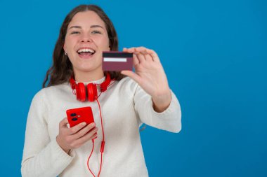 Beyaz kazaklı ve kırmızı kulaklıklı mutlu genç bir kadın kameraya bir kredi kartı gösterirken gülümsüyor ve elinde kırmızı bir akıllı telefon tutuyor, mavi arka planda, online alışverişi sembolize ediyor. Boşluğu kopyala
