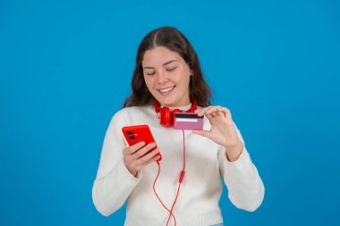Çevrimiçi ödeme yapmak için kredi kartını ve kırmızı akıllı telefonunu kullanan mutlu genç bir kadın. Boynuna kırmızı kulaklıklar takarak gülümsüyor, parlak mavi bir arka plan karşısında duruyor.. 