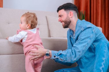 Sevgi dolu baba, pembe tulumlu, bej koltuklu, manzaralı kızı desteklerken halıya diz çöker. Nazik teşvikler, motor becerileri gelişimi ve evdeki aile bağlarını yakalar. 