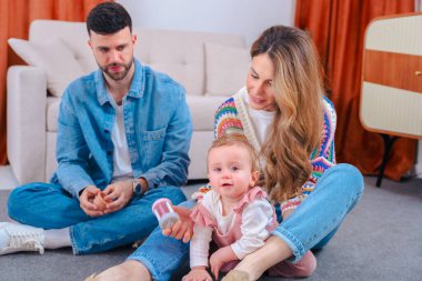 Gülümseyen ebeveynler oturma odasındaki halıda oturmuş pembe tulumlu meraklı kızlarının ahşap bir fincana uzanmasını izliyorlar. Oyuncu aile bağlarını, erken gelişimi ve ev neşesini resmediyorlar. 