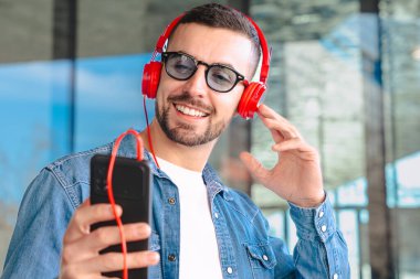 Gülümseyen beyaz adam elinde akıllı bir telefonla kırmızı kulaklıkla müzik dinlemekten hoşlanıyor. Kot gömlek giymiş, arka planda modern cam bina olan açık havada duran, parlak ve sıradan bir an.. 