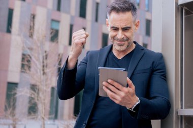 Kendine güvenen işadamı online bahsi kazanmasını kutluyor, elinde tablet, yumruğunu kaldırıyor. Şehir ortamında açık havada gülümsüyor. Başarı kavramı, dijital yaşam tarzı ve modern finans. 