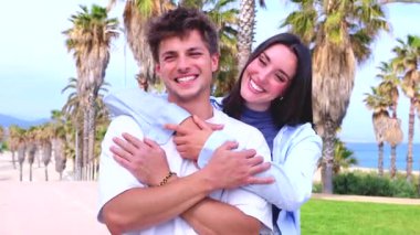 Gülümseyen genç çift, sahilin parlak bir sahil şeridinde kameraya bakarken neşeli bir kucaklaşmayı paylaşırken, romantizm, arkadaşlık ve deniz kenarındaki neşeli yaz yaşam tarzını aktarırken,. 