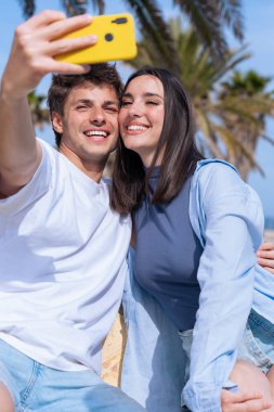 Genç bir adam ve kadın, güneşli bir avuç içi parktaki bankta oturuyor, gülümsüyor ve mutlu bir selfie çekerken, akıllı telefon, somutlaştırıcı eğlence, arkadaşlık ve yaz titreşimlerini yansıtıyorlar. Dikey