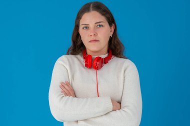 Ciddi bir ifade ve kollarını kavuşturmuş, boynunda kırmızı kulaklıklar olan genç bir kadın. Katı mavi bir arka plana karşı duruyor, memnuniyetsizliği, kararlılığı veya hoşnutsuzluğu ifade ediyor.. 