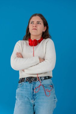 Ciddi bir ifade ve kollarını kavuşturmuş, boynunda kırmızı kulaklıklar olan genç bir kadın. Katı mavi bir arka plana karşı duruyor, memnuniyetsizliğini, kararlılığını veya hoşnutsuzluğunu ifade ediyor. Dikey