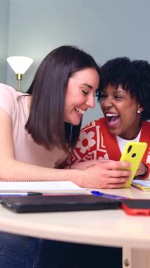  Masanın üzerine yaslanmış neşeli Afrikalı ve Kafkas arkadaşların dikey görüntüsü. Not defterleri ve kalemler arasında sarı bir akıllı telefona bakarken gülüyorlar. Çalışma molasını yakalıyorlar, internetten bağ kuruyorlar..