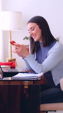 Evden çalışan neşeli genç bir kadının dikey portresi, dizüstü bilgisayar ve notlarla dolu bir masada oturuyor, kırmızı akıllı telefon mesajına gülümsüyor, uzaktan çalışmayı, bağlantıyı ve modern yaşam tarzını gösteriyor.. 