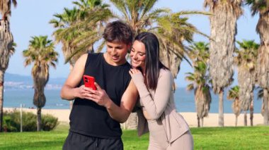 Kolsuz siyah bluzlu gülümseyen atletik adam telefonu havada tutuyor, barış işareti çakıyor, ortağı omuzuna sarılırken, hey açık gökyüzünün altındaki çimenli sahil parkında palmiye ağaçlarının arasında selfie için poz veriyor. 