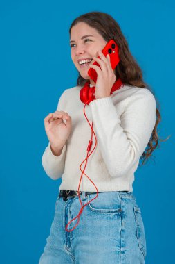 Kırmızı bir akıllı telefondan konuşan neşeli genç bir kadının dikey görüntüsü. Elini göğsüne koyup gülüyor. Parlak mavi bir arka plana yaslanıyor. Sıradan bir anda.. 