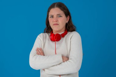 Ciddi bir ifade ve kollarını kavuşturmuş, boynunda kırmızı kulaklıklar olan genç bir kadın. Katı mavi bir arka plana karşı duruyor, memnuniyetsizliği, kararlılığı veya hoşnutsuzluğu ifade ediyor.. 