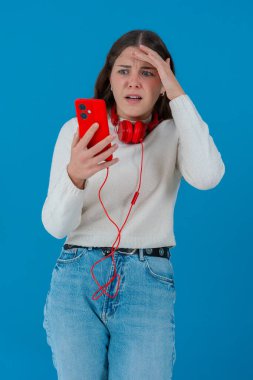 Beyaz kadın akıllı telefonundan kötü haberler okurken şok olmuş görünüyor. Kulaklık takıyor ve beyaz bir kazak giyiyor. Katı mavi arka plana karşı kaygıyla tepki veriyor..