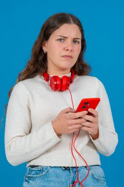 Genç bir kadın kırmızı bir akıllı telefon tutarken kafası karışmış ve hayal kırıklığına uğramış görünüyor. Boynuna kırmızı kulaklıklar takarak parlak mavi bir arka plan karşısında bir soruna veya hataya tepki veriyor. Dikey