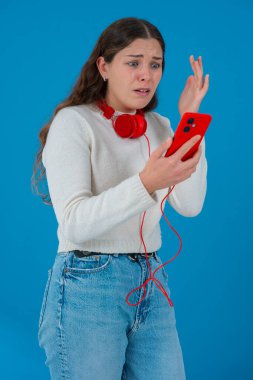 Beyaz kadın akıllı telefonundan kötü haberler okurken şok olmuş görünüyor. Kulaklık takıyor ve beyaz bir kazak giyiyor. Katı mavi arka plana karşı kaygıyla tepki veriyor..