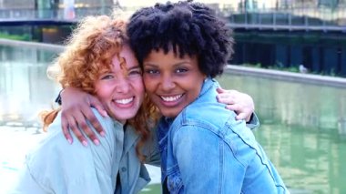  Afrika kökenli Amerikalı ve Kafkasyalı dostlar modern bir şehir kanalının yanında kucaklaşıyorlar, kameraya doğru gülümsüyorlar, arkadaşlığı, çeşitliliği ve dışarıda neşeli anları kutluyorlar. 